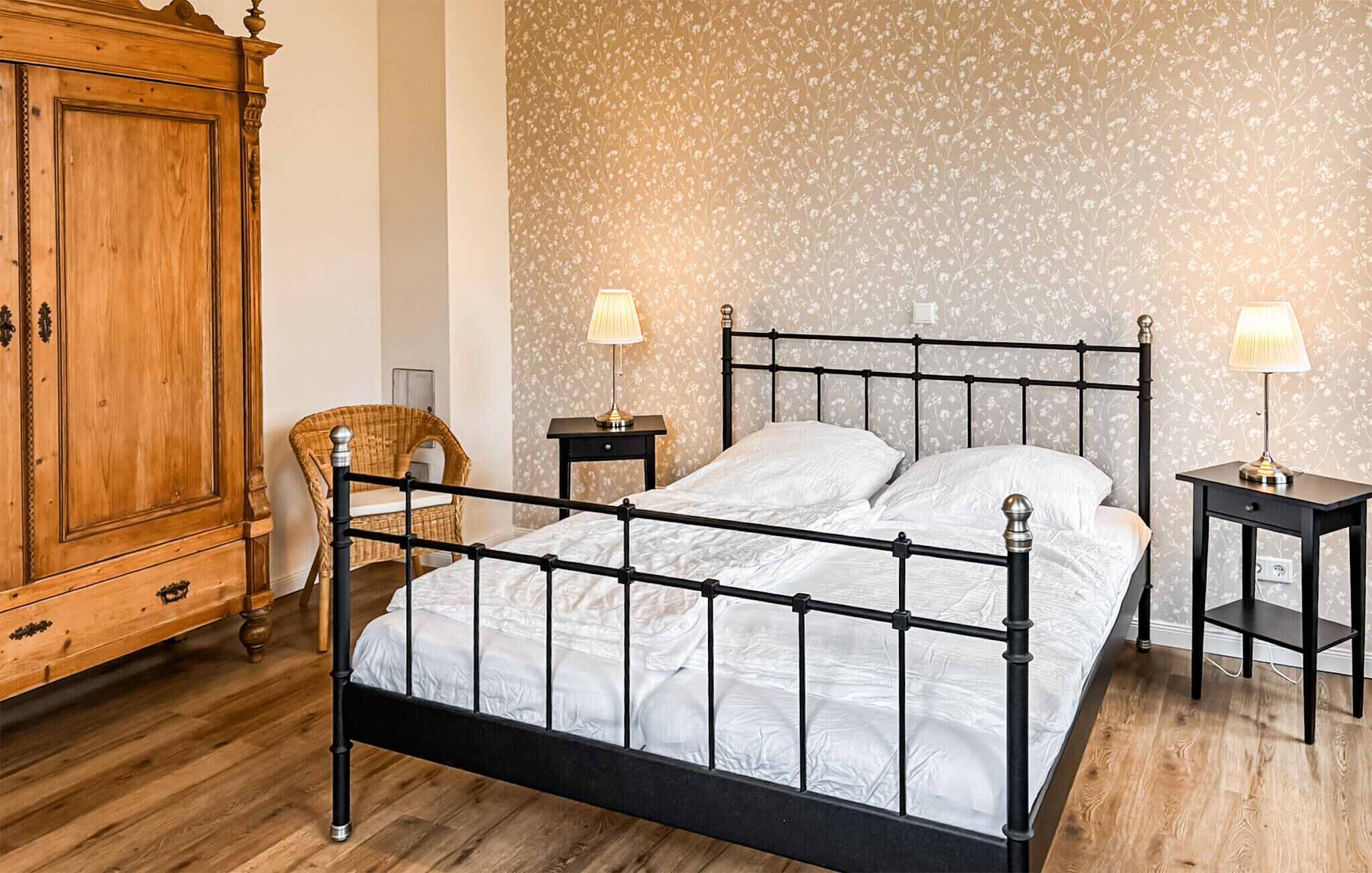 A bedroom with a metal bed frame, wooden wardrobe, wicker chair, two bedside tables with lamps, and floral wallpaper.