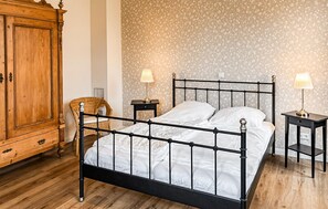 Ein Schlafzimmer mit einem Metallbettgestell, einem Holzschrank, einem Korbstuhl, zwei Nachttischen mit Lampen und blumigem Wandpapier.