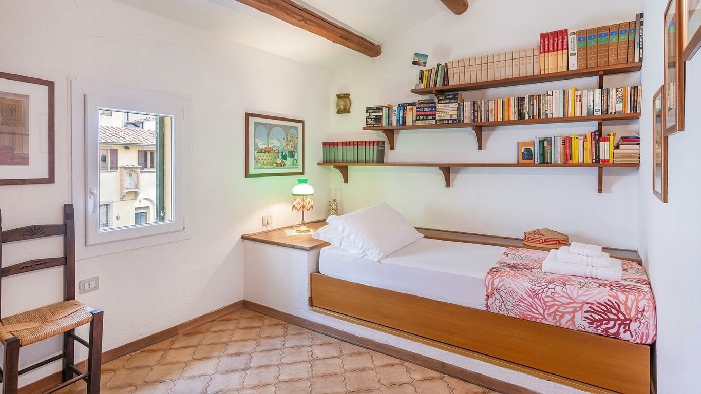 A bedroom with a bed, a nightstand, a lamp, a chair, a window, and bookshelves.