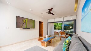 A modern living room with a flat-screen TV, a ceiling fan, a dining area with a glass table, and a large window with a view of greenery. - Big Terrace Luxury 2-bedroom condo in charming Playa del Carmen with AC (Playa del Carmen)