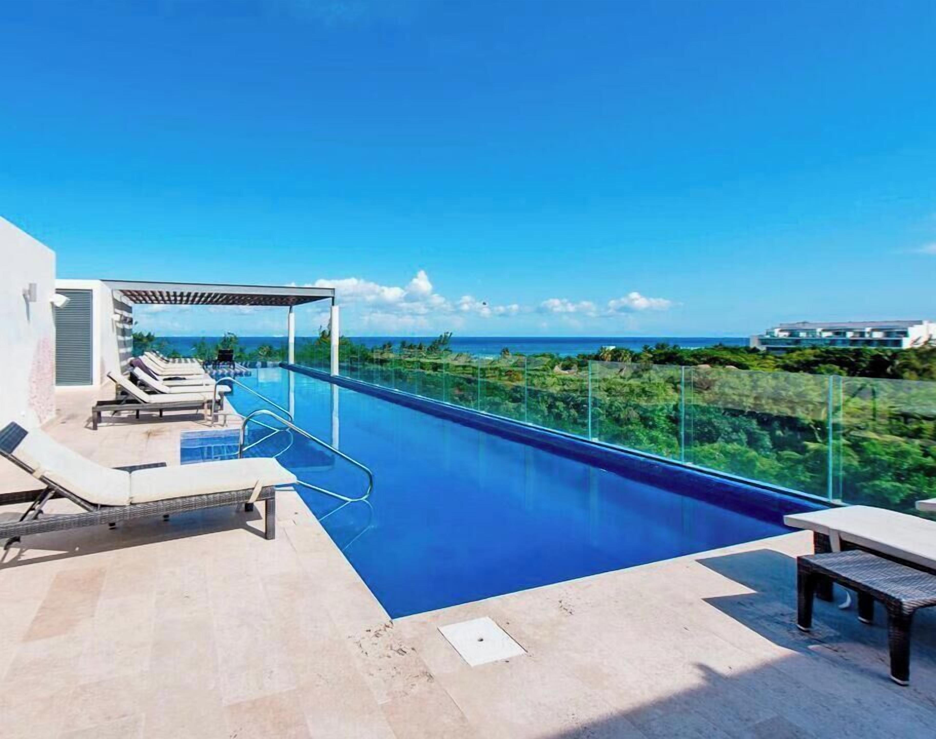 A clear blue pool with lounge chairs and a covered seating area, overlooking a lush landscape.