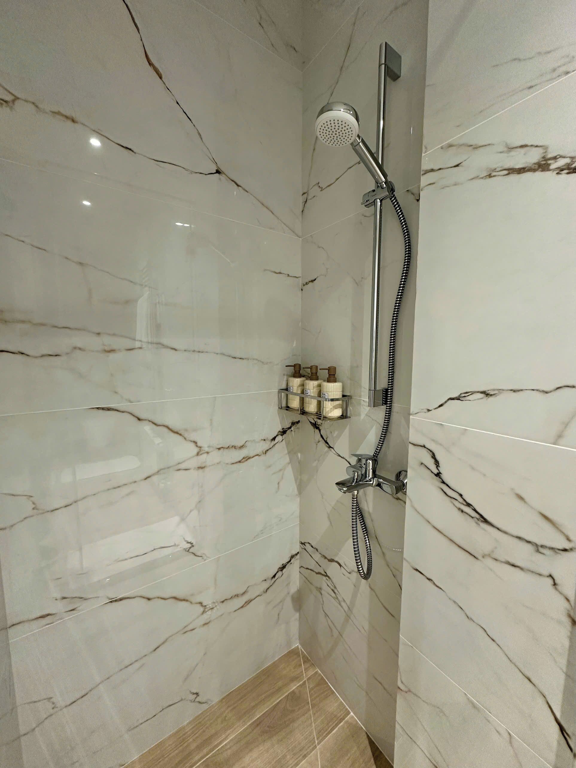 Un baño con paredes de mármol, ducha con cabezal de ducha manual y repisa para jabón.