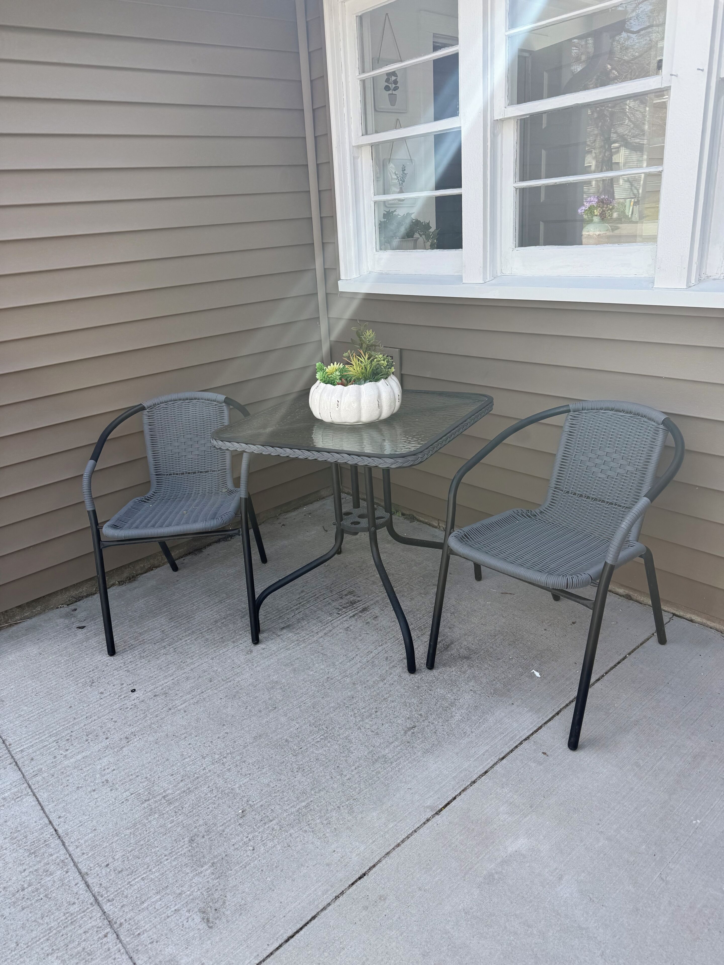 Un pequeño patio al aire libre con una mesa de vidrio y dos sillas de mimbre, una planta en maceta sobre la mesa y una ventana con persianas.
