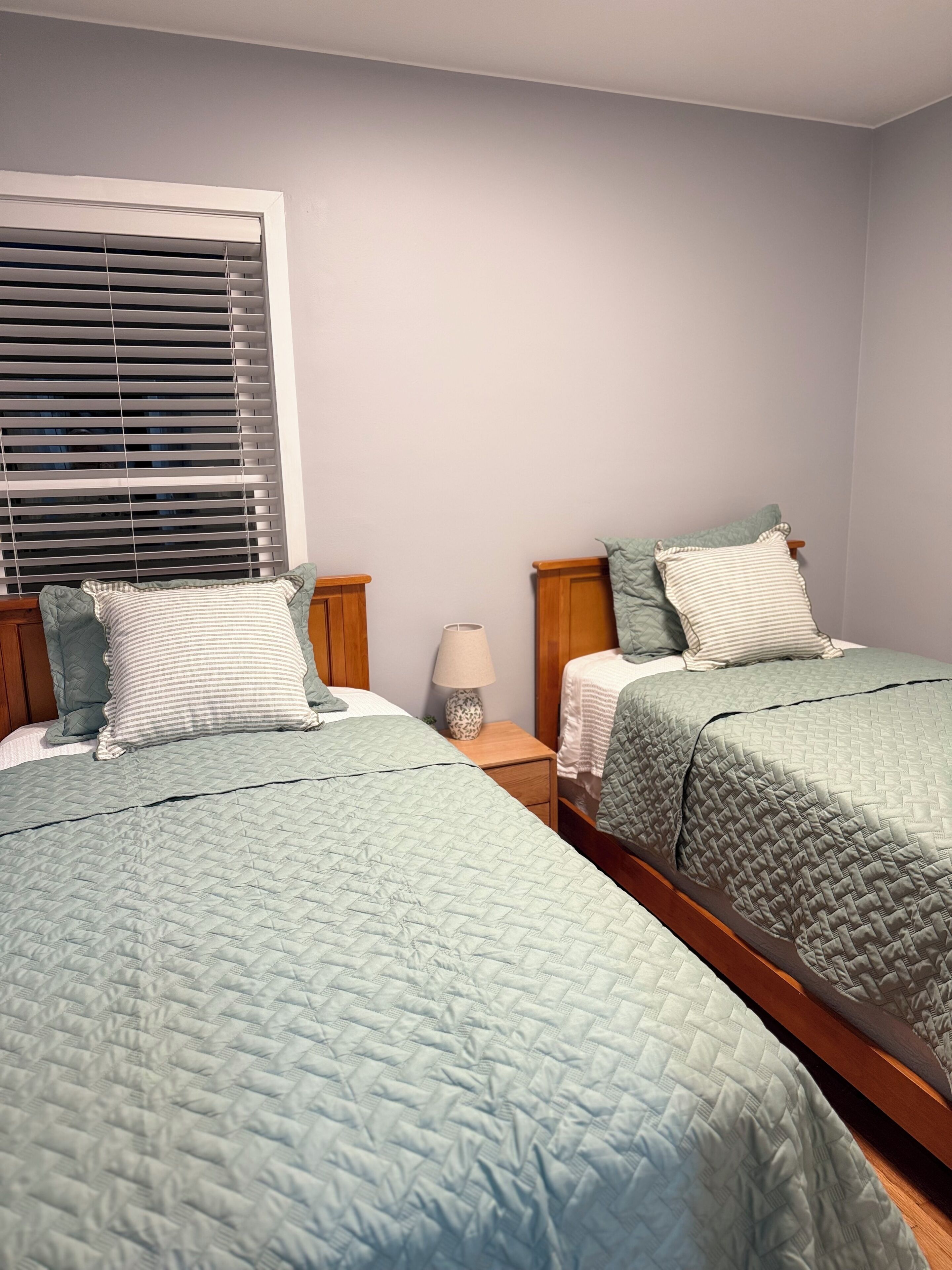 Un dormitorio con dos camas, una ventana con persianas, una mesita de noche con una lámpara y una lámpara de pared.
