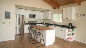 A modern kitchen with white cabinets, stainless steel appliances, a wooden island with bar stools, and a window providing natural light. - Waterfront Fishing Lodge | 2BR | Dockside Charters (Ketchikan)