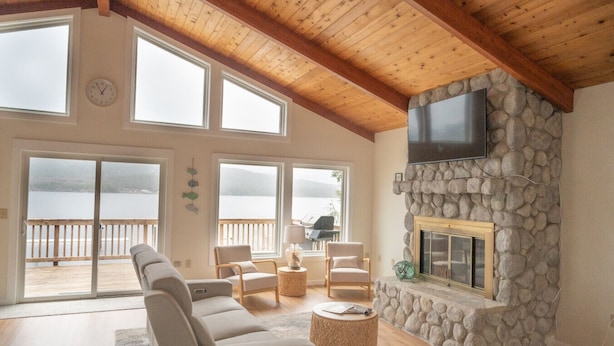 A spacious living room with a stone fireplace, large windows, and a wooden ceiling. - Waterfront Fishing Lodge | 2BR | Dockside Charters (Ketchikan)