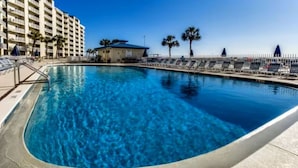 Una piscina con agua cristalina azul, rodeada por una zona de piscina con sillones de descanso y un edificio al fondo.