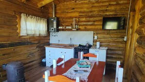 A wooden cabin interior with a kitchen area, a dining table with chairs, a stove, a sink, and a television mounted on the wall.