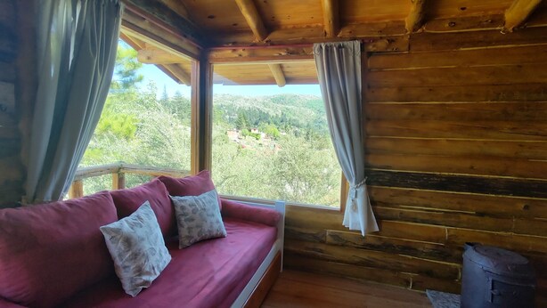 A wooden cabin interior with a sofa, curtains, a view of greenery, and a wood stove.