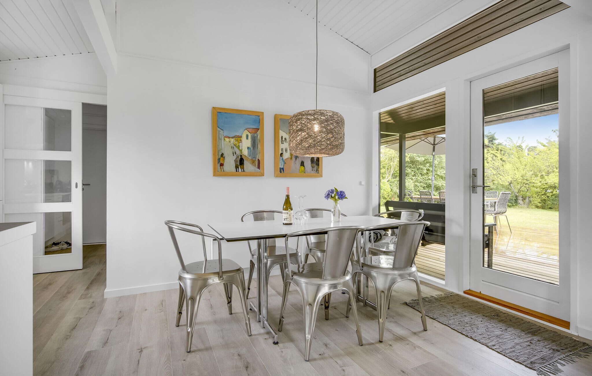 Un espace repas avec une table blanche, des chaises en métal, deux tableaux, un luminaire suspendu et une vue sur l’extérieur.