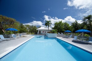 Piscina con lettini e ombrelloni blu, circondata da alberi e un edificio con tetto bianco.