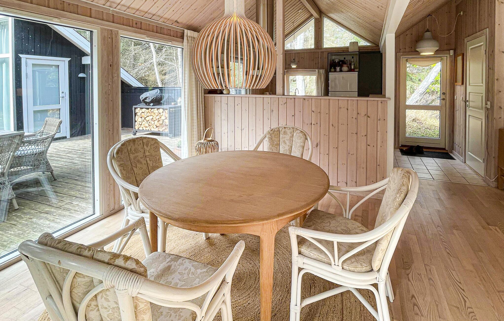 Un intérieur en bois avec une table ronde et des chaises en osier, un coin cuisine et de grandes fenêtres.