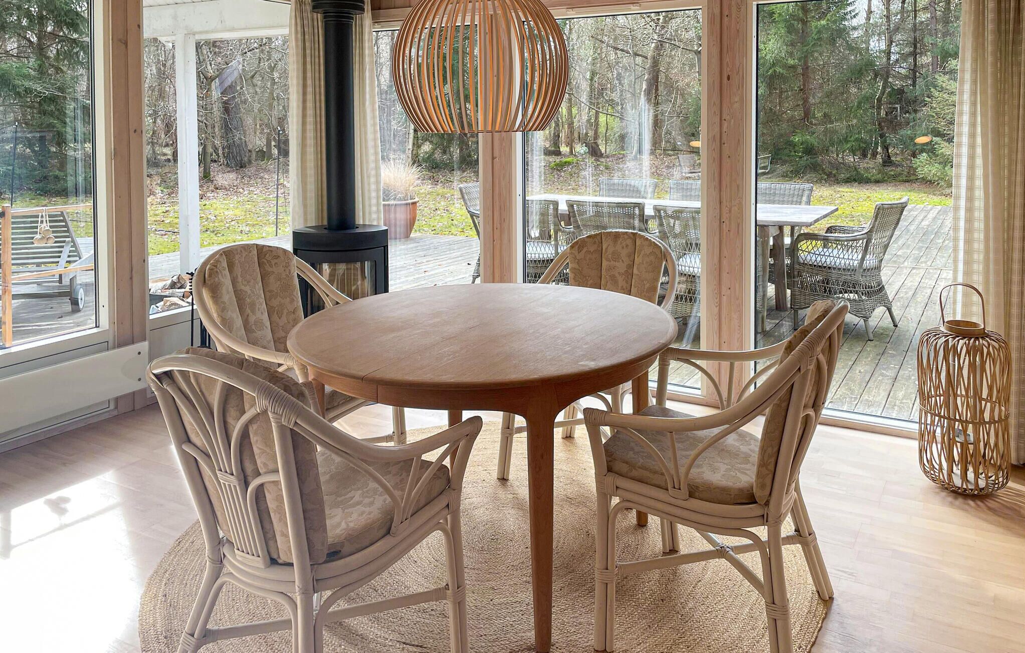 A round wooden table with four wicker chairs, a hanging light fixture, and a view of a deck with outdoor furniture.