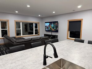Una sala de estar moderna con un televisor grande, un sofá empotrado, una isla de cocina con fregadero y ventanas amplias.