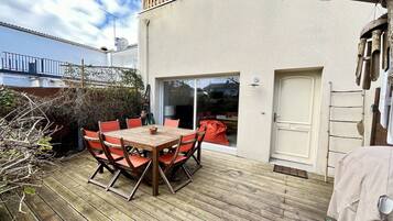 Una terraza de madera con una mesa, sillas, un ventanal corredero y una puerta blanca.