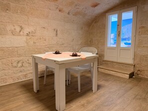 A room with a stone wall, a white table with a pink cloth, a wicker chair, and a view through a door. - Casa Antonia by Monholiday (Monopoli)