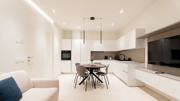 Una cocina moderna con armarios blancos, una mesa de comedor con sillas, un sofá con un cojín azul y un televisor de pantalla plana grande montado en la pared.