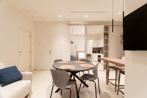 A modern living room with a round dining table, grey chairs, a sofa with a blue pillow, and a kitchen in the background. - Solymar Mare by Monholiday (Monopoli)