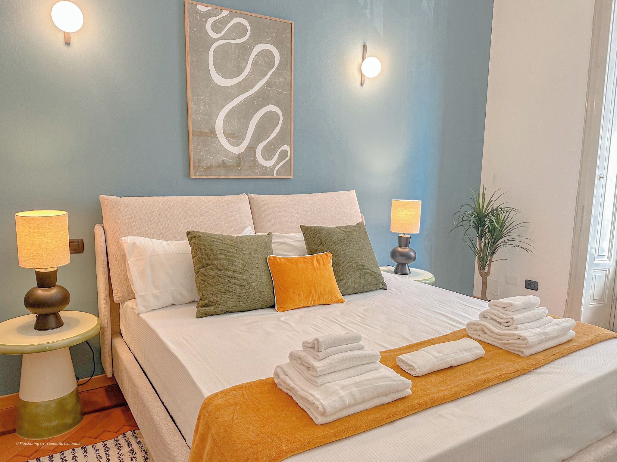 A neatly made bed with white linens, two side tables with lamps, a wall-mounted light fixture, and a framed abstract artwork above the bed.