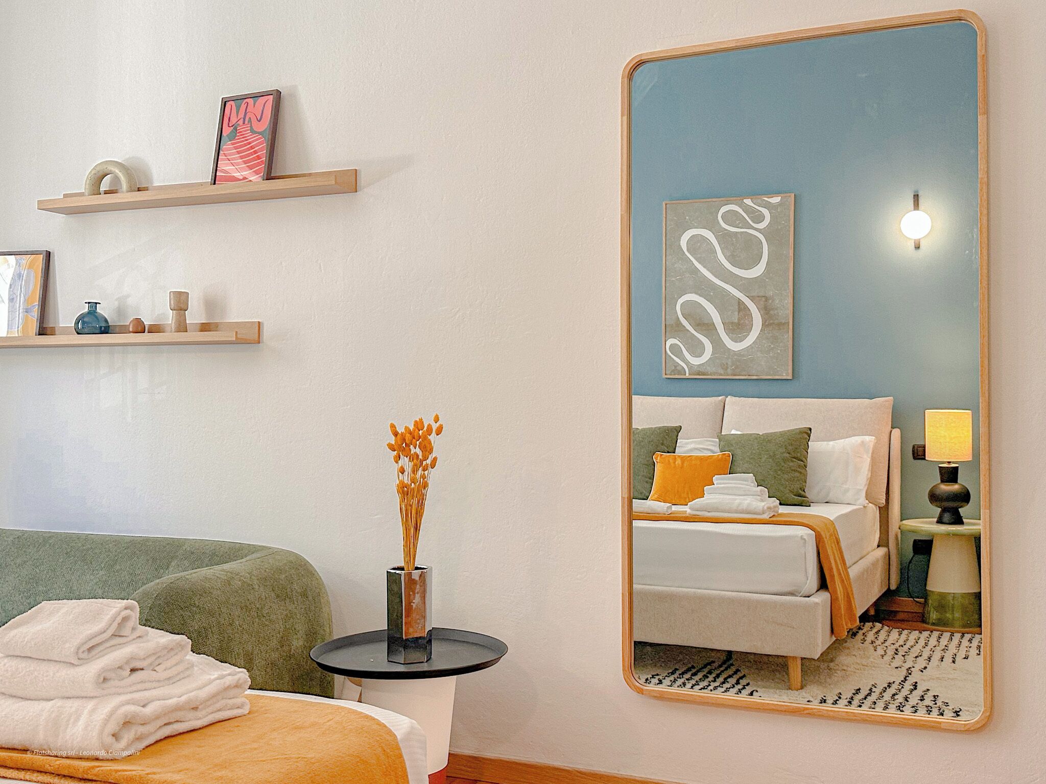 Une chambre d’hôtel comprenant un lit, une table de chevet avec un vase, un miroir, une étagère avec des objets décoratifs et un mur orné d’une œuvre d’art encadrée.