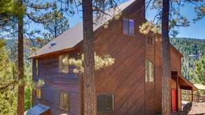Una cabaña de madera con techo inclinado, ventanas amplias y una puerta roja, rodeada de árboles y un paisaje montañoso.