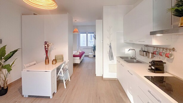 Una cucina moderna con mobili bianchi, un'area pranzo con tavolo e sedie bianche e un corridoio che conduce a una camera da letto.