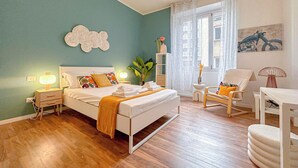 A bedroom with a bed, bedside tables, a chair, a small table, a lamp, and a window with curtains. - Flatsharing - Gioia Blue Retreat – 3 Bedrooms (Milan)