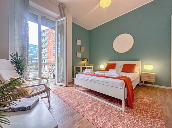 Quarto com cama, mesa de cabeceira, abajur, janela com cortinas e vista para edifícios.
