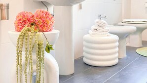 Une salle de bain avec un lavabo, des toilettes et un bidet, tous de couleur blanche. On peut y voir des fleurs roses sur le lavabo et une pile de serviettes blanches.