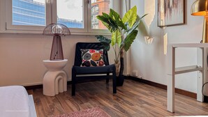 Une pièce avec une grande fenêtre, une plante en pot, une chaise avec un coussin, une petite table et une lampe.
