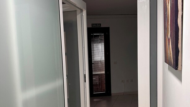 Ein Flur mit einer Tür, auf der 'SALIDA' steht, die zu einem Raum mit einem Fenster und Jalousien führt.