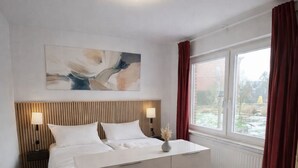 Ein Schlafzimmer mit einem Bett, Nachttischen, einer Wandleuchte, einem Fenster mit Vorhängen und Blick ins Freie.