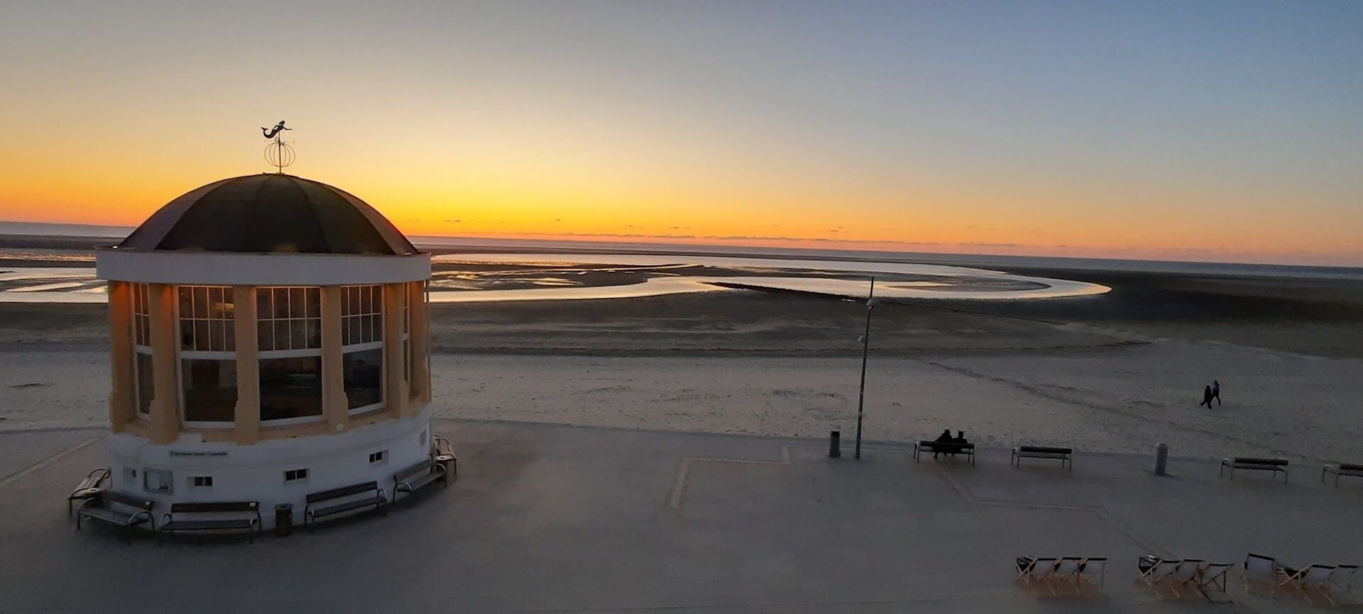 Ein Gebäude mit Kuppel und Wetterfahne, umgeben von einer Sandfläche mit Bänken und blauem Himmel.