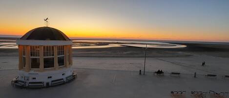 Ein Gebäude mit Kuppel und Wetterfahne, umgeben von einer Sandfläche mit Bänken und blauem Himmel.