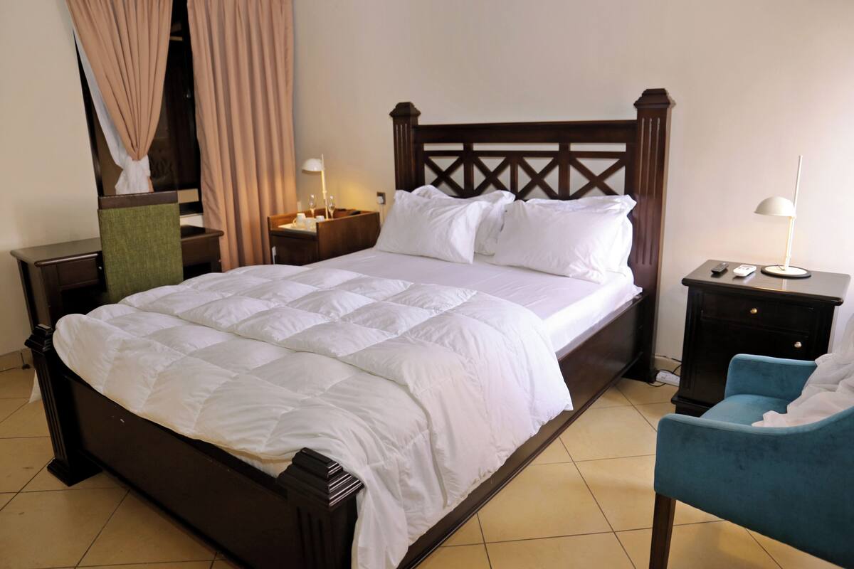 A neatly made bed with white linens, a wooden headboard, a bedside table with a lamp, and a chair.