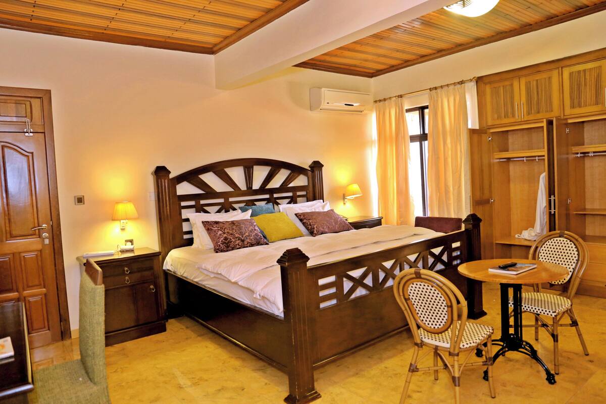 A wooden four-poster bed with pillows, a bedside table, a chair, a small round table, and a wardrobe.