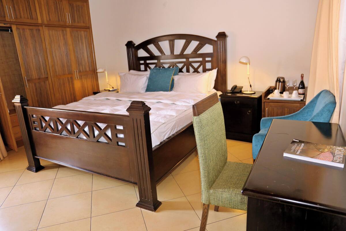 A hotel room with a wooden bed, a green upholstered chair, a small table with a magazine, and a nightstand with a lamp and a phone.