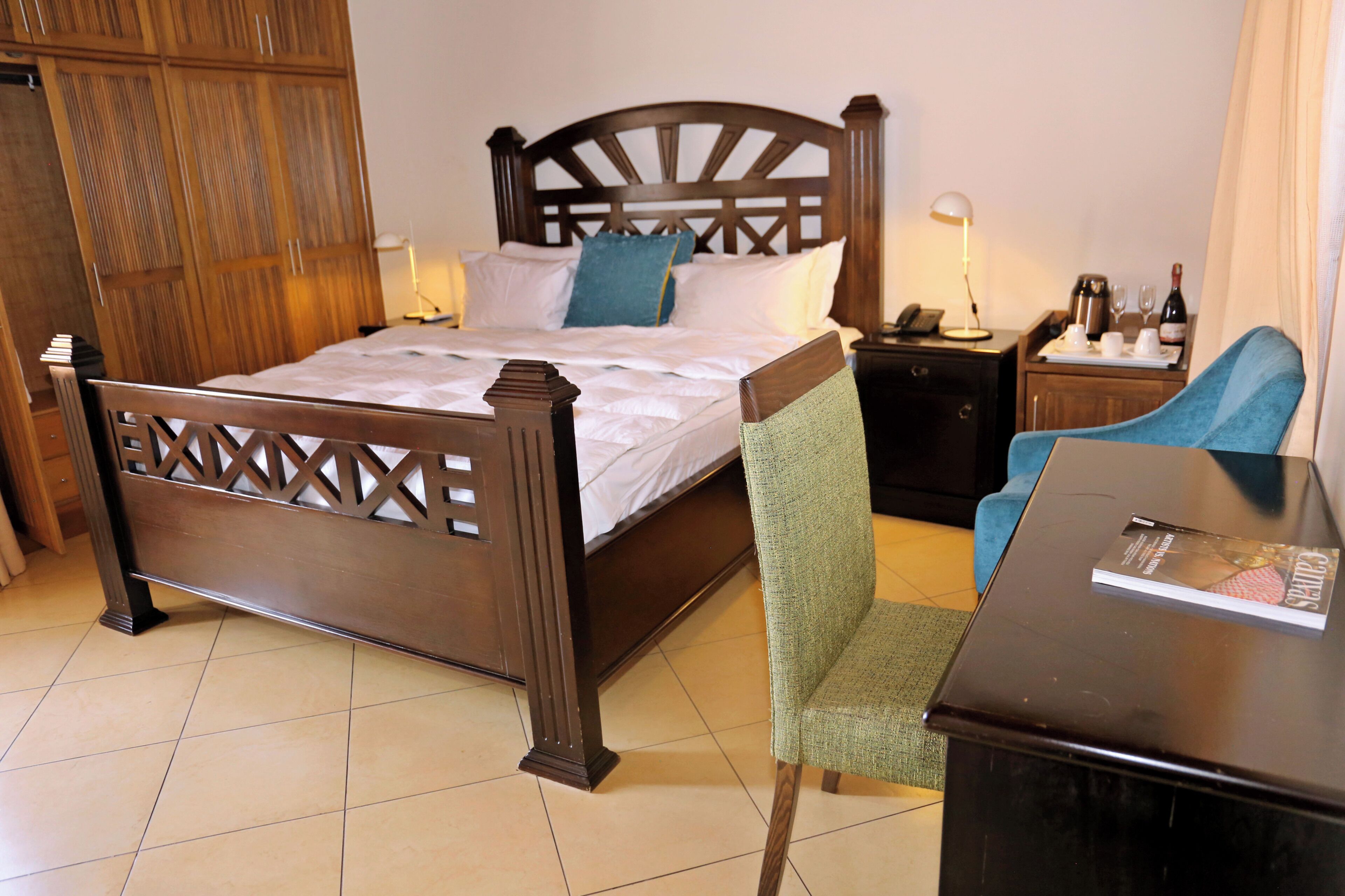 A hotel room with a wooden bed, a green chair, a desk with a magazine, and a nightstand with a lamp and a phone.