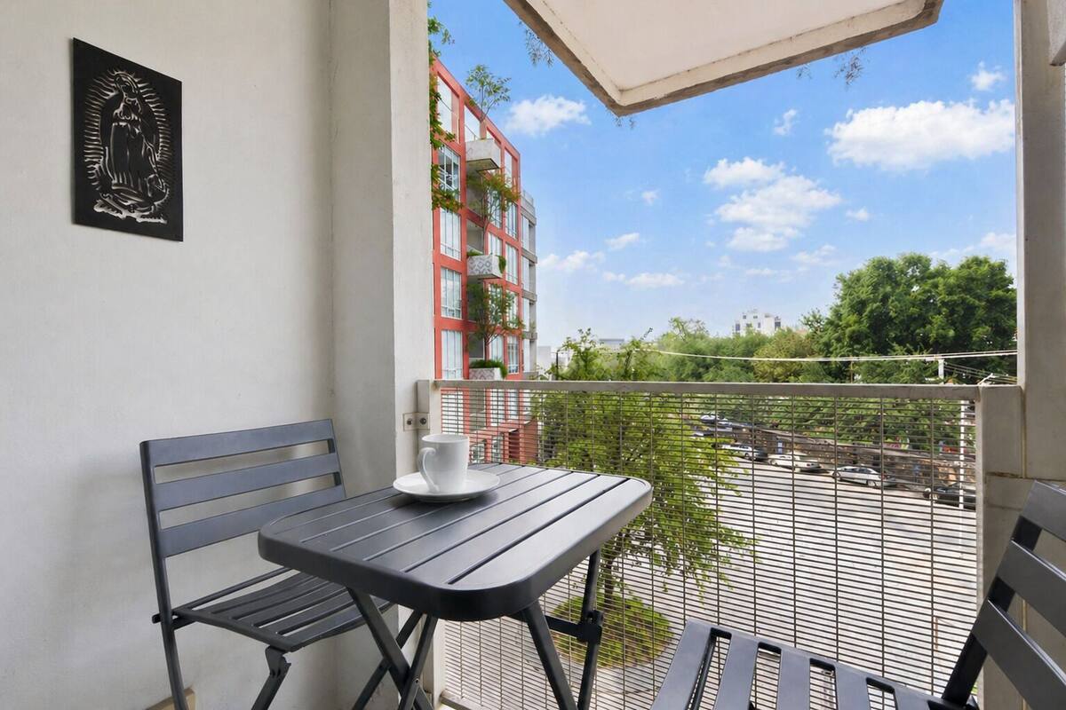 Un balcone con tavolo e sedie, un'opera d'arte incorniciata e vista su un edificio rosso e aree verdi.