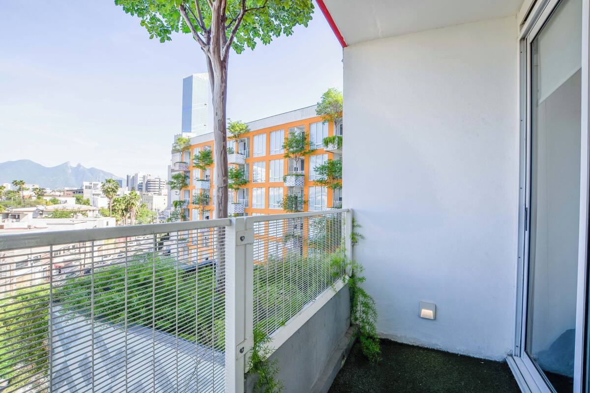 Un balcone con ringhiera in metallo, un albero e vista su un paesaggio urbano con edifici e montagne.
