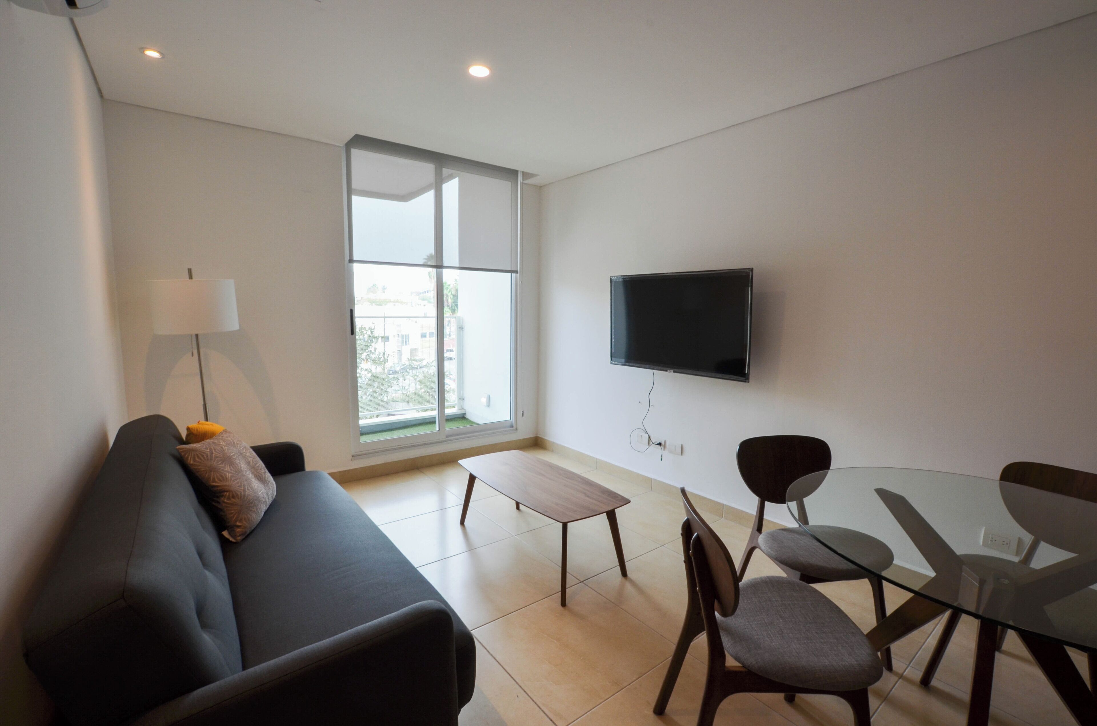 Een moderne woonkamer met een bank, een glazen tafel en stoelen, een flatscreen televisie aan de muur en een groot raam voor natuurlijk licht.