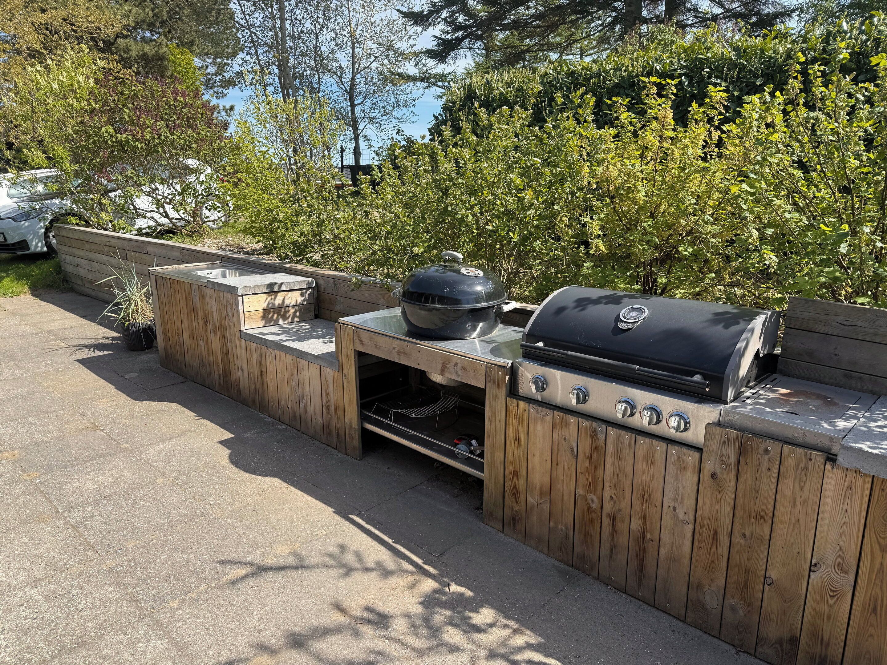 Un área de barbacoa exterior de madera con una parrilla y un lavadero, rodeada de follaje.