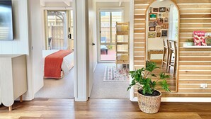 Ein Zimmer mit Holzboden, einem Bett mit roter Decke, einem Topfpflanzen, einem Spiegel mit Holzrahmen und einem Regal mit verschiedenen Gegenständen.
