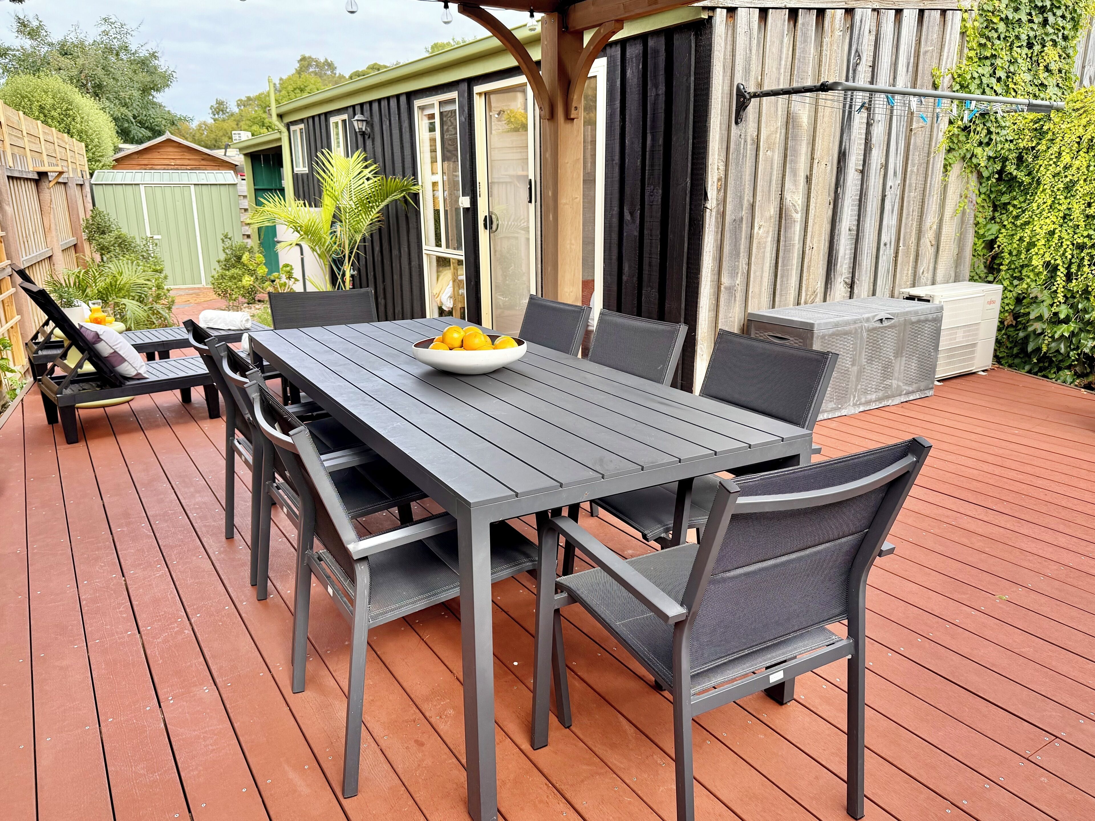 Une terrasse extérieure couverte, avec une table et des chaises, un saladier de fruits et, au fond, une clôture en bois.