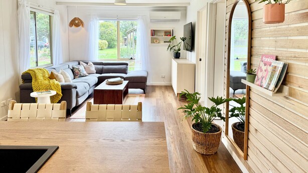 Ein modernes Wohnzimmer mit einem Ecksofa, einem Couchtisch und einem an der Wand montierten Fernseher. Üchtüpfungen und ein Bücherregal mit Büchern und Dekorationsobjekten sind ebenfalls vorhanden.