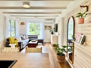 A modern living room with a sectional sofa, a coffee table, and a TV mounted on the wall. There are potted plants and a bookshelf with books and decorative items. - The Salty Grove - A Retro Coastal Cottage (Ocean Grove)