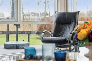 Una habitación moderna con una butaca de cuero negro, una mesa de vidrio con adornos, y vistas a árboles a través de amplios ventanales.