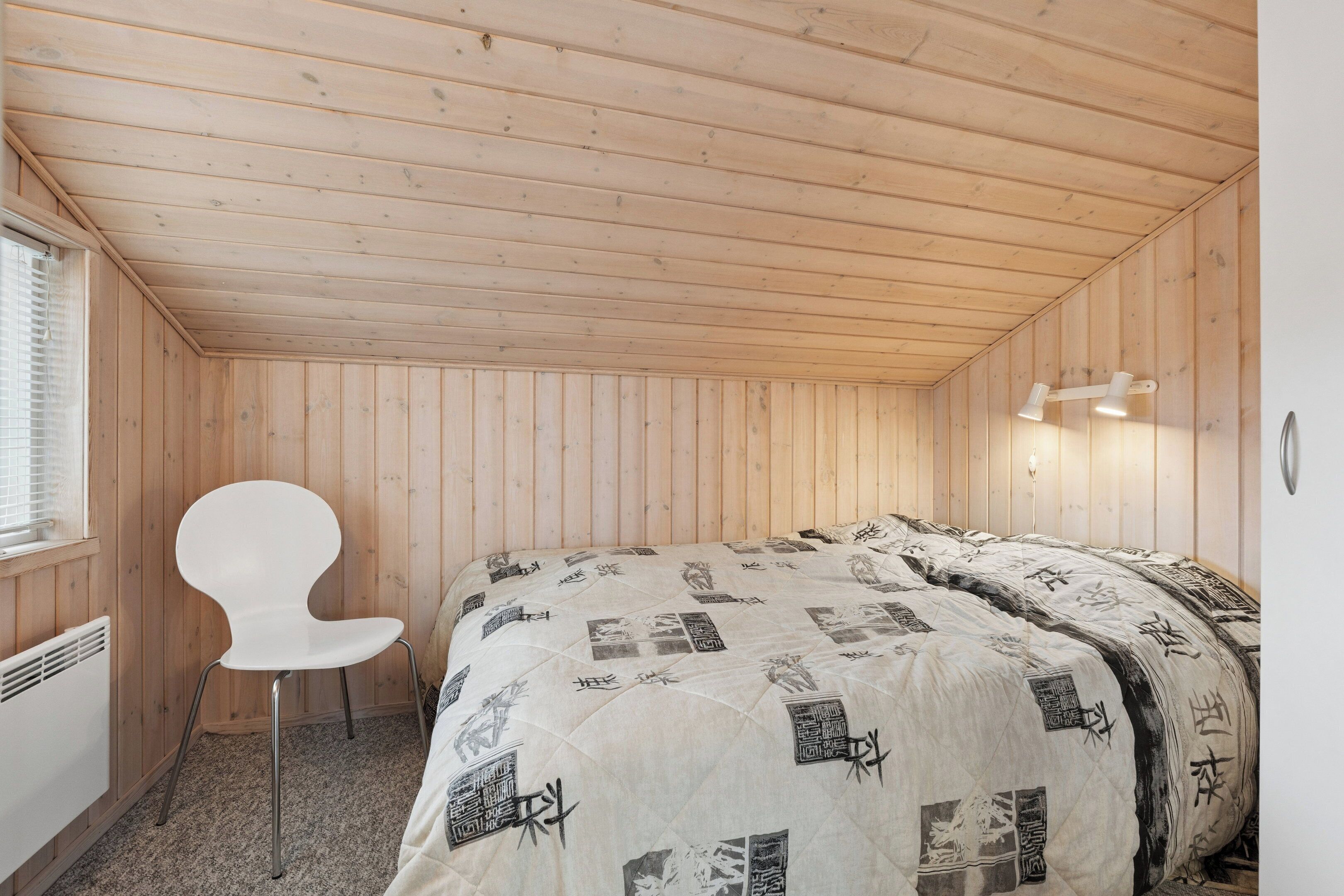 Una habitación con paneles de madera, una cama, una silla blanca y una lámpara fijada a la pared.