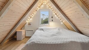 Ett sovrum med ett sluttande tak, en säng med ett geometriskt mönstrat överkast, ett litet sängbord, en nattlampa, ett fönster med utsikt över grönskan och ljusslingor.