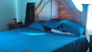 A bedroom with a wooden bed, a nightstand, and a window with curtains. - Welcome to Grace Guest House. A peaceful and comfortable home. (St. Elizabeth Parish)